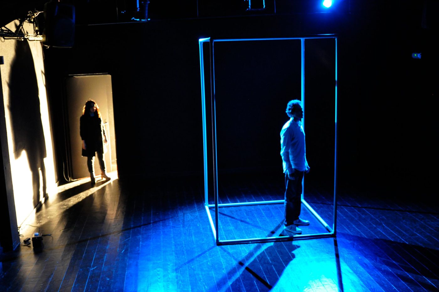 Escena de Apenas 20 segundos: un hombre dentro de una estructura iluminada en azul y una mujer en una puerta al fondo